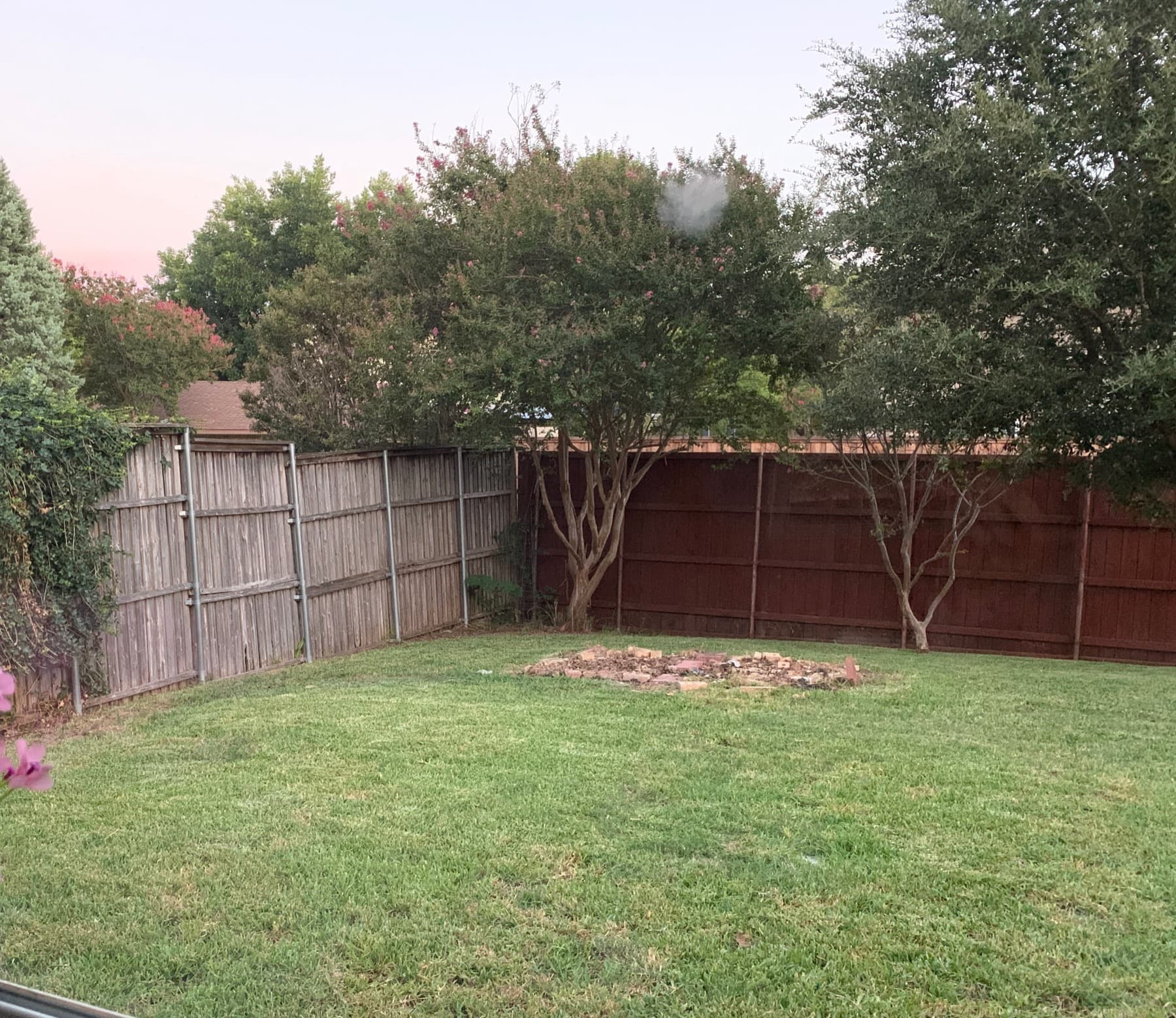 An empty back lawn with a weathered fence and a bare fire-pit ring